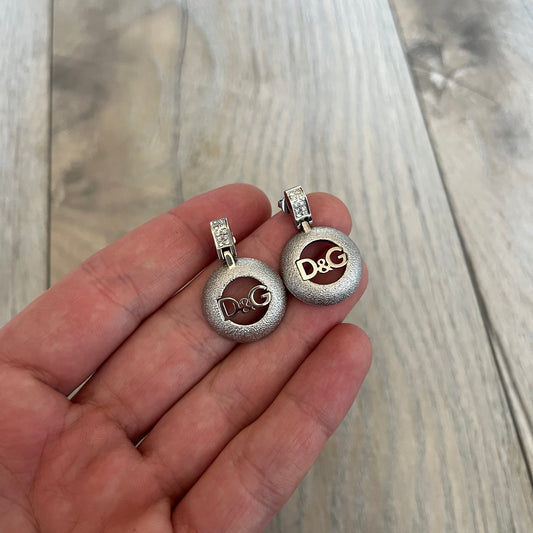 Dolce & Gabbana 2000s Round D&G Logo Earrings feature a dangling design with thick 3D circular charms shaped with a cut-out center and D&G logo in the middle. The connector between the charm and earring post is embellished with crystal detailing. The earrings are finished in a silver-tone metal construction.