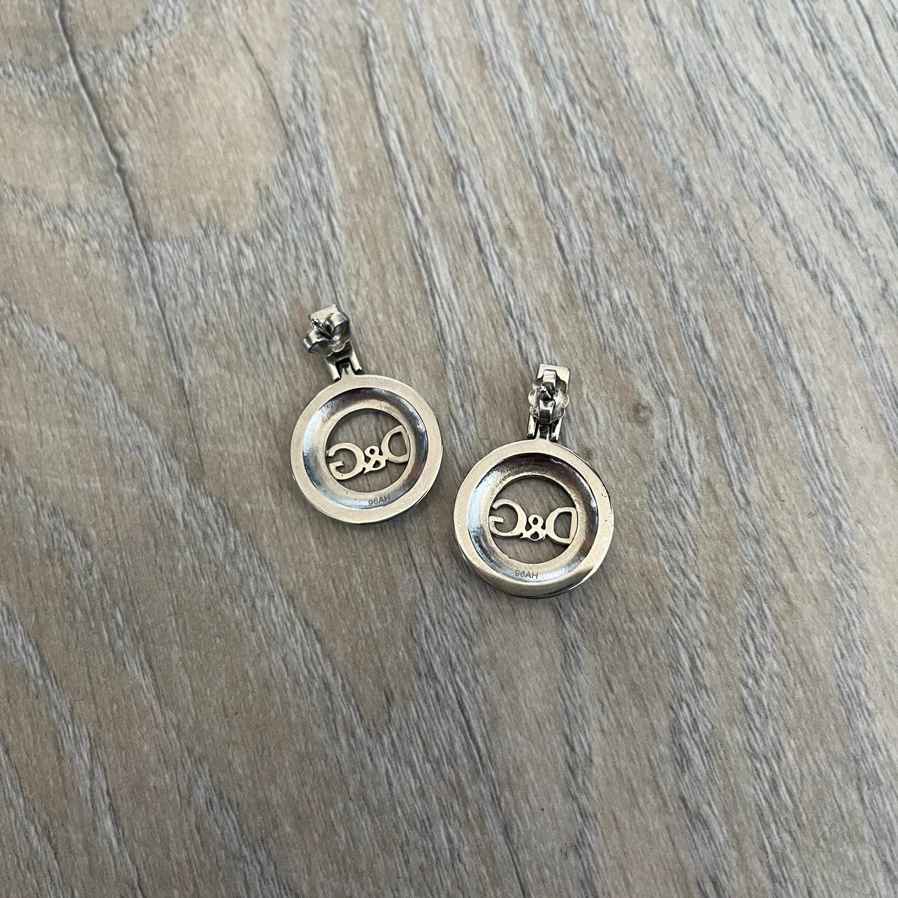Dolce & Gabbana 2000s Round D&G Logo Earrings feature a dangling design with thick 3D circular charms shaped with a cut-out center and D&G logo in the middle. The connector between the charm and earring post is embellished with crystal detailing. The earrings are finished in a silver-tone metal construction.