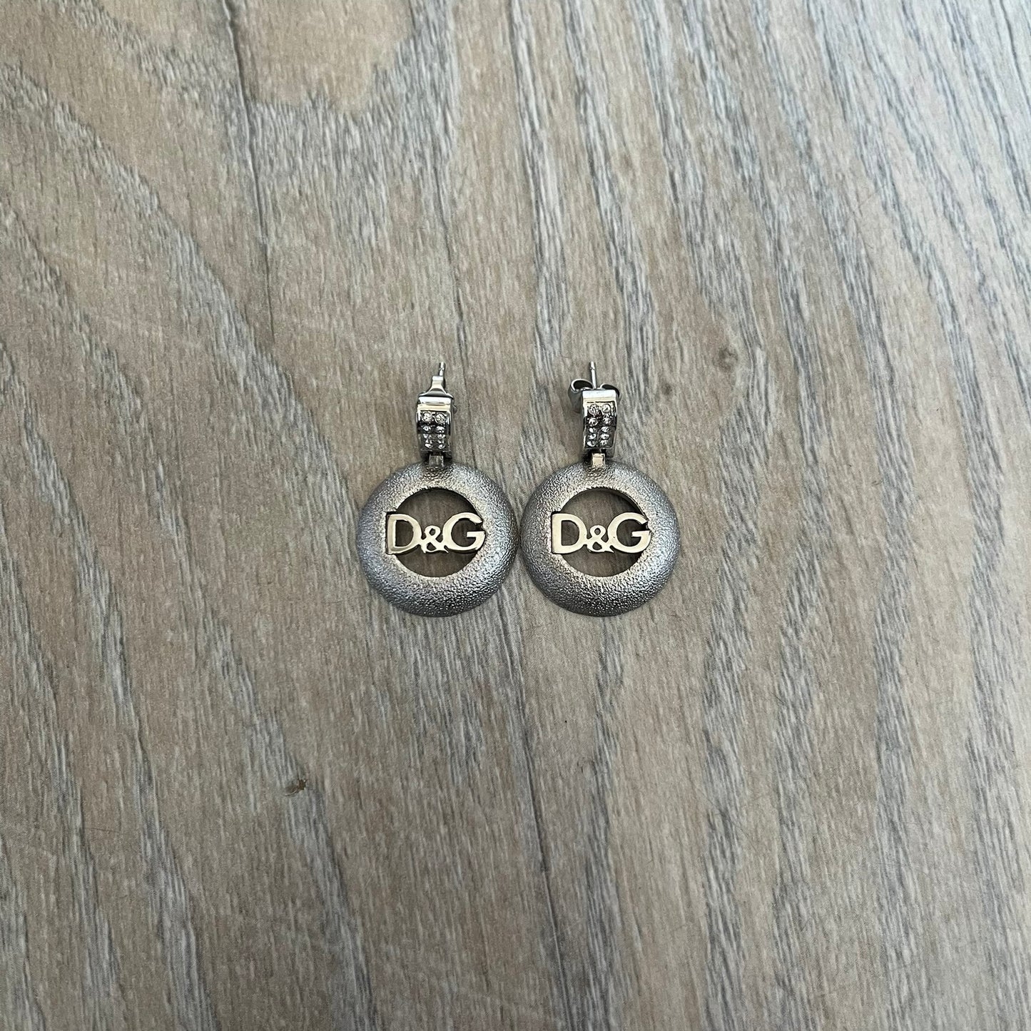 Dolce & Gabbana 2000s Round D&G Logo Earrings feature a dangling design with thick 3D circular charms shaped with a cut-out center and D&G logo in the middle. The connector between the charm and earring post is embellished with crystal detailing. The earrings are finished in a silver-tone metal construction.