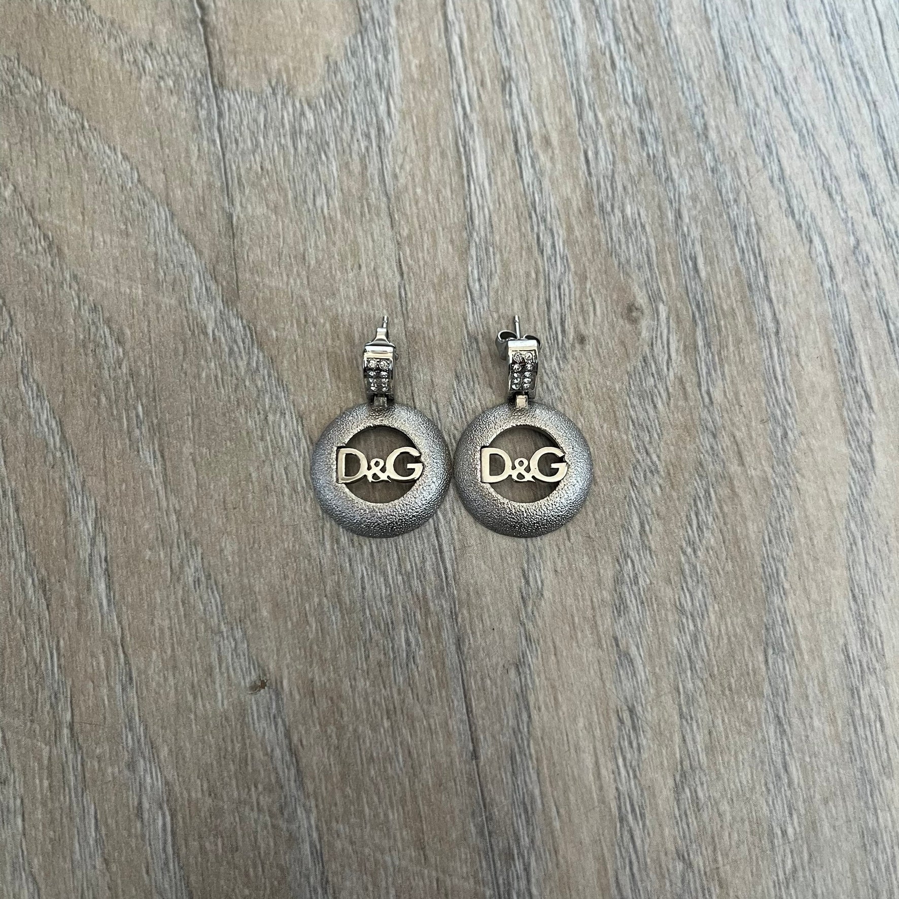 Dolce & Gabbana 2000s Round D&G Logo Earrings feature a dangling design with thick 3D circular charms shaped with a cut-out center and D&G logo in the middle. The connector between the charm and earring post is embellished with crystal detailing. The earrings are finished in a silver-tone metal construction.