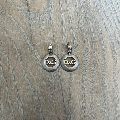 Dolce & Gabbana 2000s Round D&G Logo Earrings feature a dangling design with thick 3D circular charms shaped with a cut-out center and D&G logo in the middle. The connector between the charm and earring post is embellished with crystal detailing. The earrings are finished in a silver-tone metal construction.