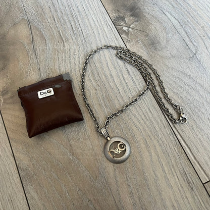 Dolce & Gabbana 2000s Round D&G Logo Necklace is a chain necklace featuring a lobster clasp closure and a thick 3D circular pendant with cut-out center and D&G logo in the middle. The pendant connector is embellished with crystal detailing. The necklace comes with a brown Dolce & Gabbana dust bag.