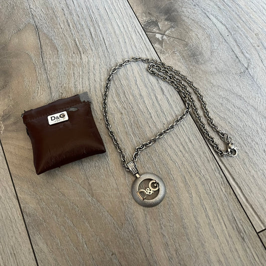 Dolce & Gabbana 2000s Round D&G Logo Necklace is a chain necklace featuring a lobster clasp closure and a thick 3D circular pendant with cut-out center and D&G logo in the middle. The pendant connector is embellished with crystal detailing. The necklace comes with a brown Dolce & Gabbana dust bag.