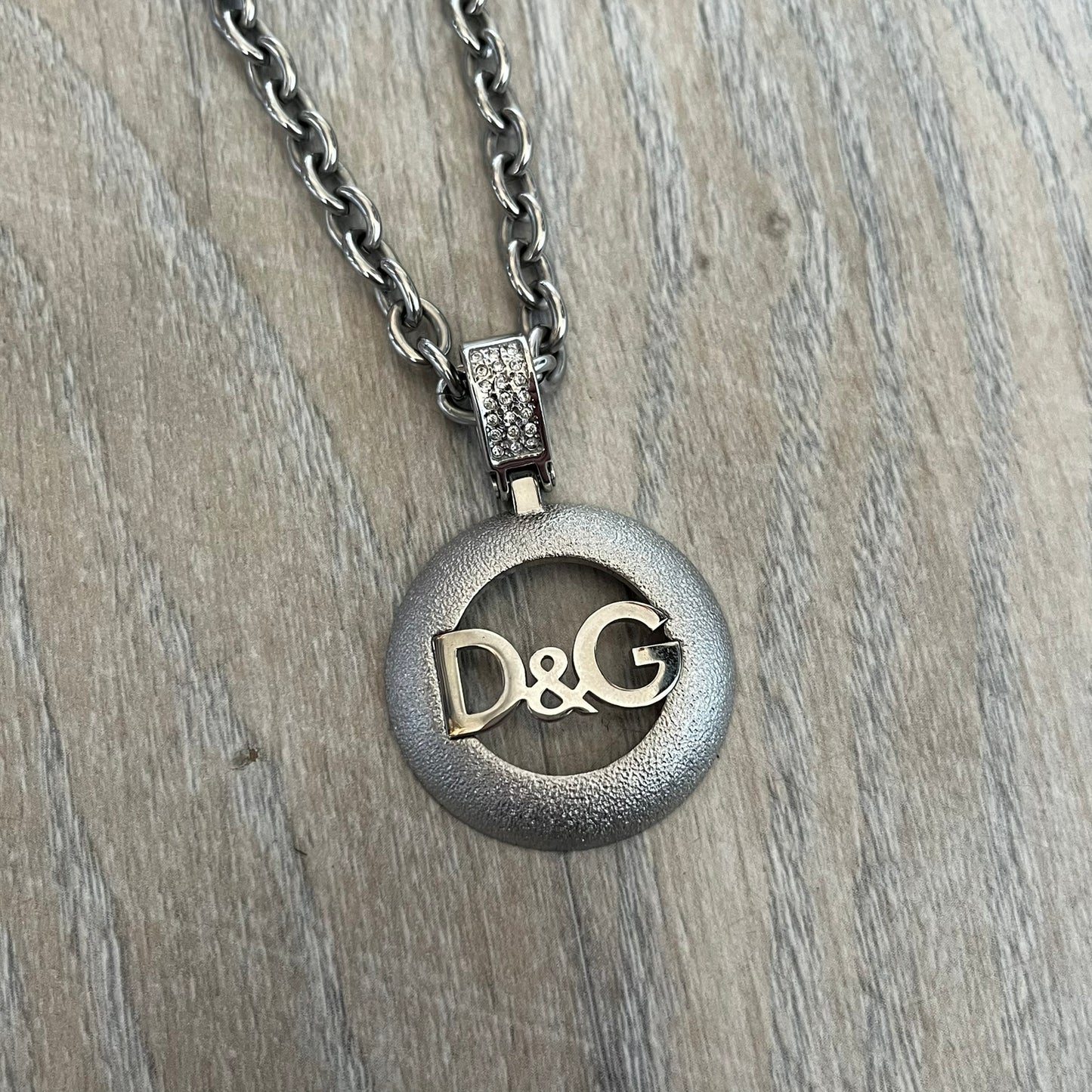 Dolce & Gabbana 2000s Round D&G Logo Necklace is a chain necklace featuring a lobster clasp closure and a thick 3D circular pendant with cut-out center and D&G logo in the middle. The pendant connector is embellished with crystal detailing. The necklace comes with a brown Dolce & Gabbana dust bag.