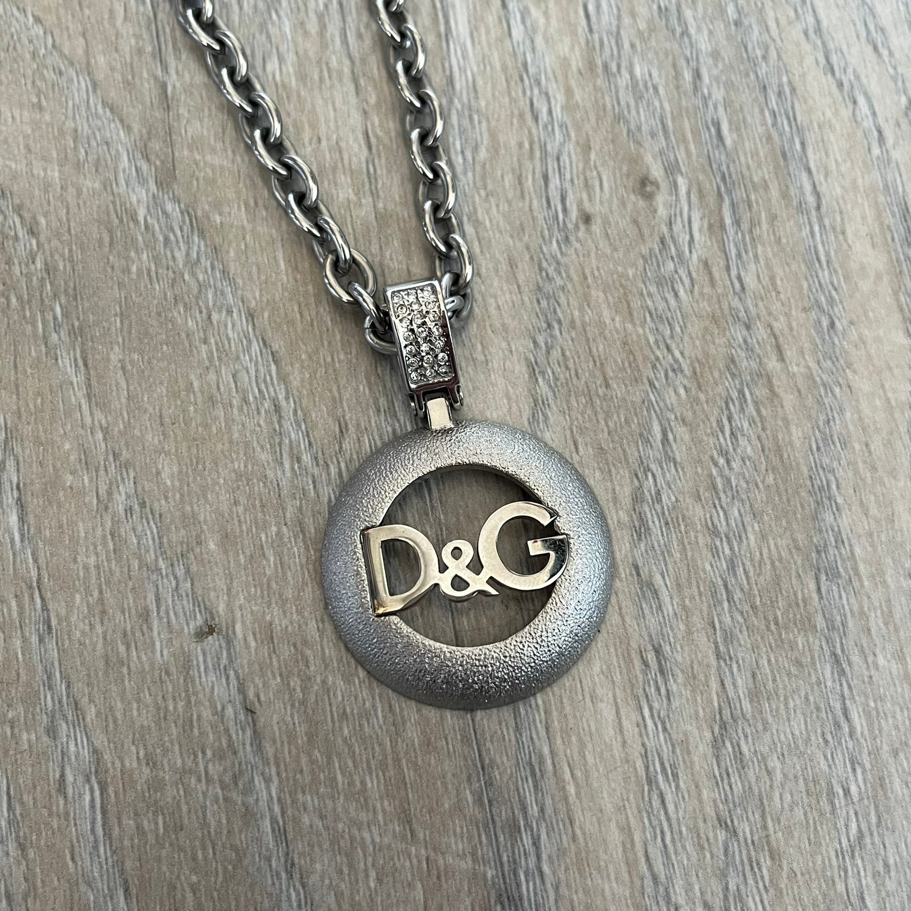 Dolce & Gabbana 2000s Round D&G Logo Necklace is a chain necklace featuring a lobster clasp closure and a thick 3D circular pendant with cut-out center and D&G logo in the middle. The pendant connector is embellished with crystal detailing. The necklace comes with a brown Dolce & Gabbana dust bag.