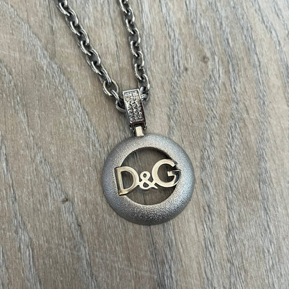 Dolce & Gabbana 2000s Round D&G Logo Necklace is a chain necklace featuring a lobster clasp closure and a thick 3D circular pendant with cut-out center and D&G logo in the middle. The pendant connector is embellished with crystal detailing. The necklace comes with a brown Dolce & Gabbana dust bag.