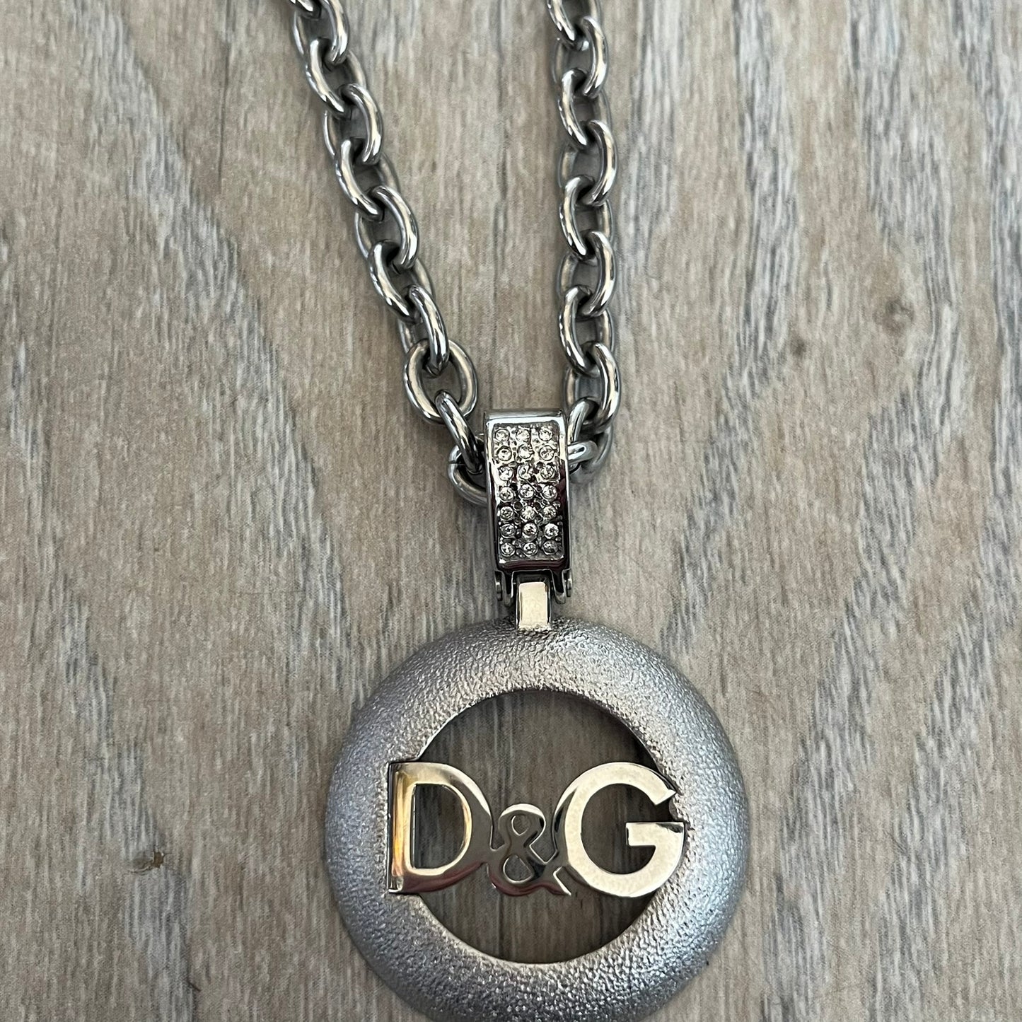 Dolce & Gabbana 2000s Round D&G Logo Necklace is a chain necklace featuring a lobster clasp closure and a thick 3D circular pendant with cut-out center and D&G logo in the middle. The pendant connector is embellished with crystal detailing. The necklace comes with a brown Dolce & Gabbana dust bag.