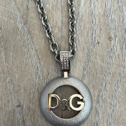 Dolce & Gabbana 2000s Round D&G Logo Necklace is a chain necklace featuring a lobster clasp closure and a thick 3D circular pendant with cut-out center and D&G logo in the middle. The pendant connector is embellished with crystal detailing. The necklace comes with a brown Dolce & Gabbana dust bag.