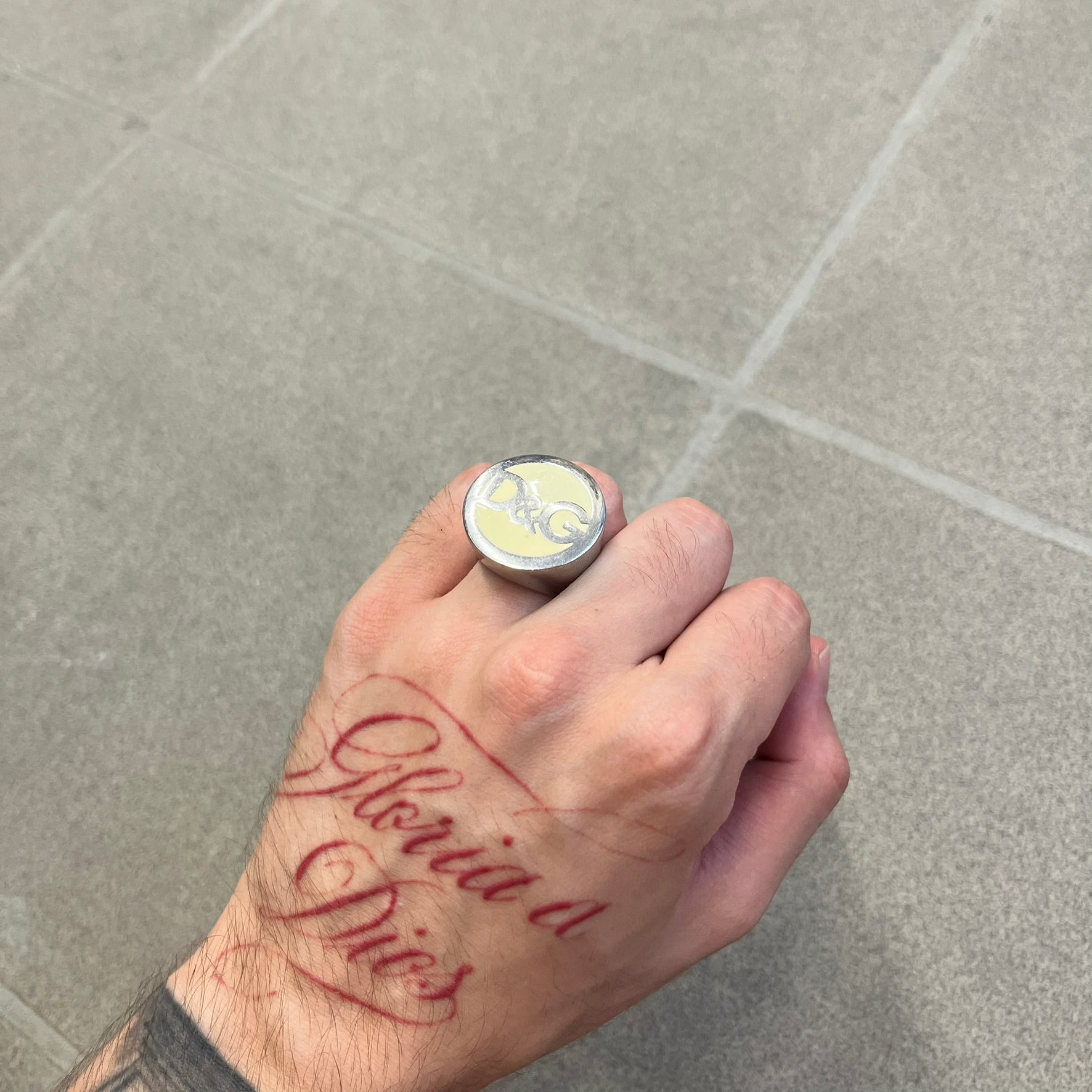 The Dolce &amp; Gabbana Silver Logo Signet Ring is a silver-tone metal signet ring with a thick, substantial design. The top features a beige background with the D&amp;G logo in silver tone, outlined in silver. The signet is large and solid.