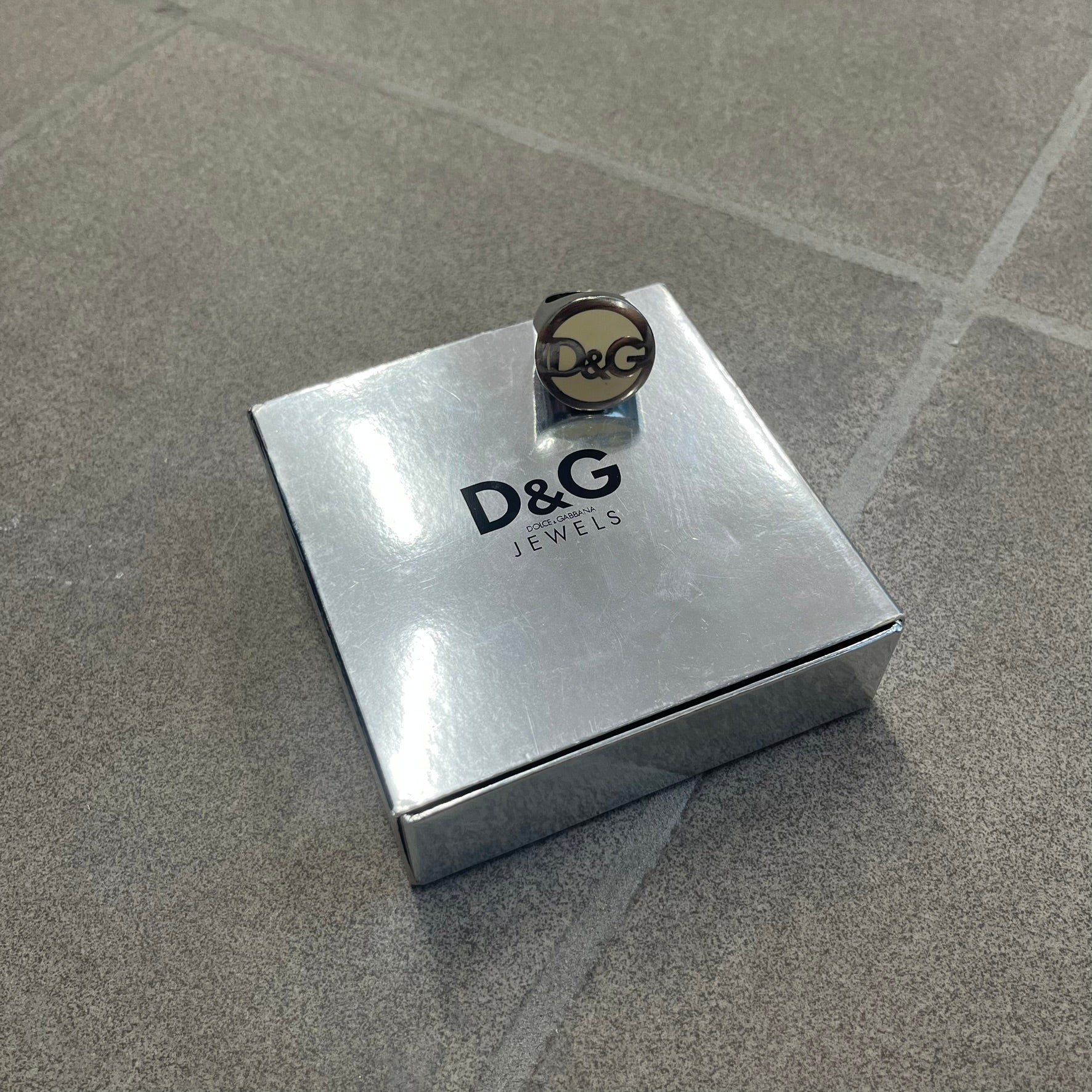 The Dolce &amp; Gabbana Silver Logo Signet Ring is a silver-tone metal signet ring with a thick, substantial design. The top features a beige background with the D&amp;G logo in silver tone, outlined in silver. The signet is large and solid.