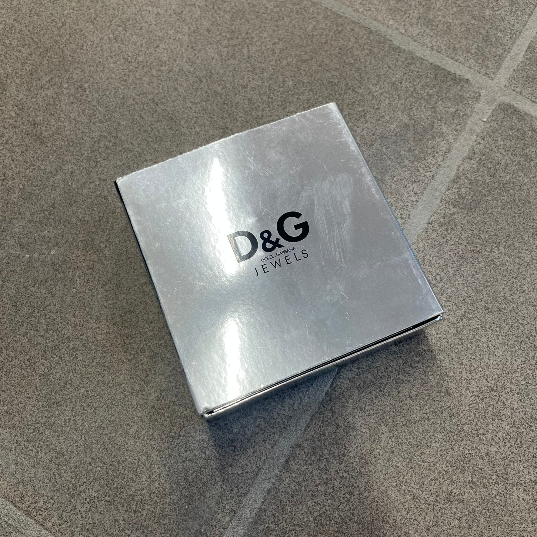 The Dolce &amp; Gabbana Silver Logo Signet Ring is a silver-tone metal signet ring with a thick, substantial design. The top features a beige background with the D&amp;G logo in silver tone, outlined in silver. The signet is large and solid.
