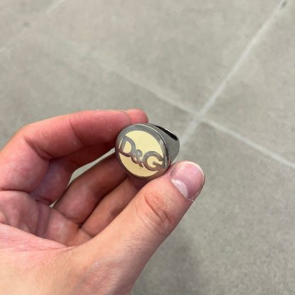 The Dolce &amp; Gabbana Silver Logo Signet Ring is a silver-tone metal signet ring with a thick, substantial design. The top features a beige background with the D&amp;G logo in silver tone, outlined in silver. The signet is large and solid.