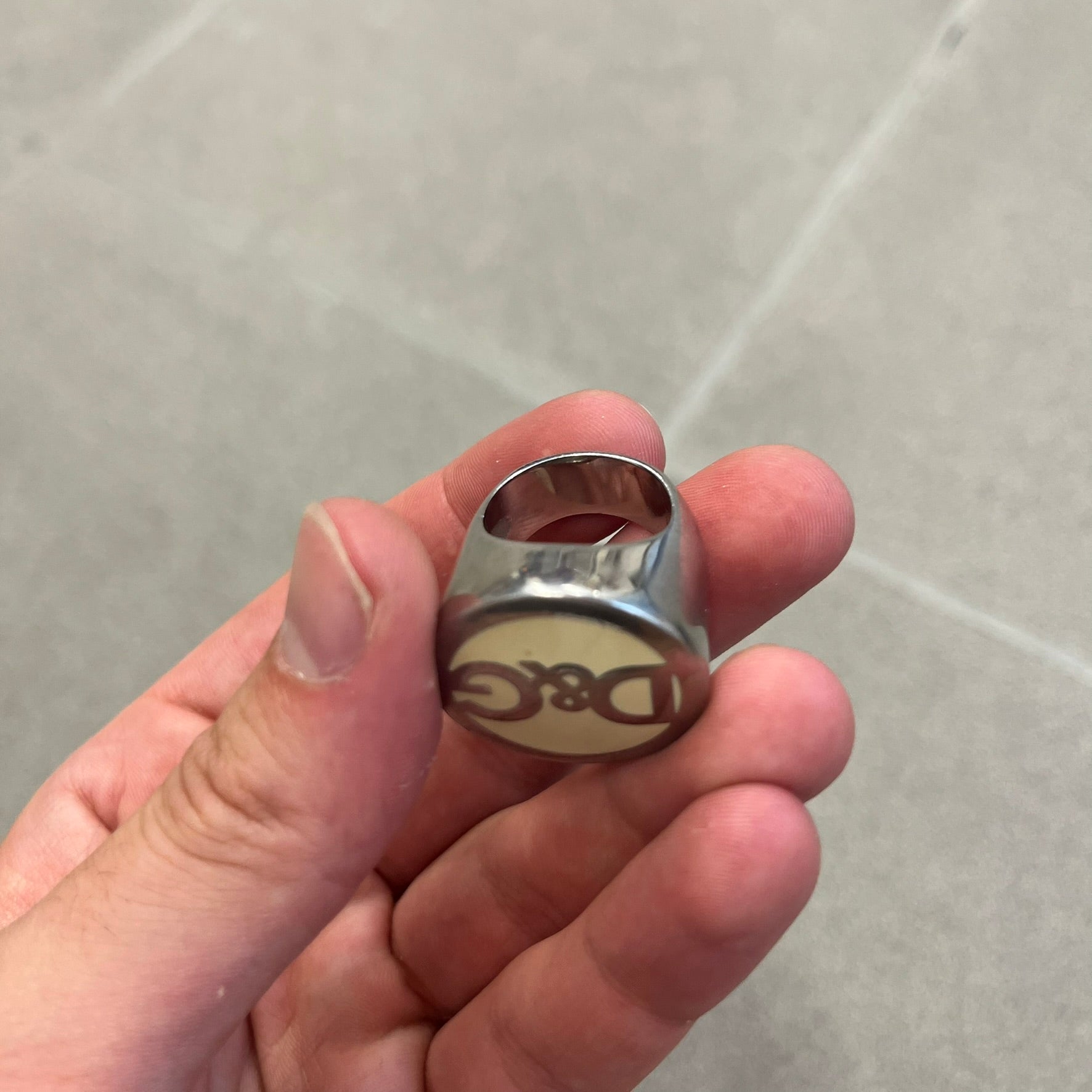 The Dolce &amp; Gabbana Silver Logo Signet Ring is a silver-tone metal signet ring with a thick, substantial design. The top features a beige background with the D&amp;G logo in silver tone, outlined in silver. The signet is large and solid.