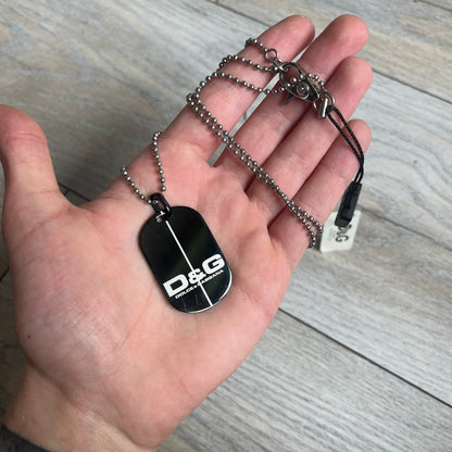 Dolce & Gabbana 2000s Split Black Dog Tag Necklace is a stainless steel necklace featuring a black dog tag pendant with a vertical grey split line running from top to bottom. The lower section of the pendant includes D&G and Dolce & Gabbana logos in the same grey tone as the split detail. The pendant has an iridescent black finish that shifts color depending on the angle. It is attached to a steel ball chain with a lobster clasp closure.