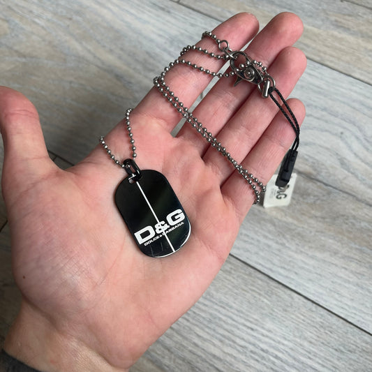 Dolce & Gabbana 2000s Split Black Dog Tag Necklace is a stainless steel necklace featuring a black dog tag pendant with a vertical grey split line running from top to bottom. The lower section of the pendant includes D&G and Dolce & Gabbana logos in the same grey tone as the split detail. The pendant has an iridescent black finish that shifts color depending on the angle. It is attached to a steel ball chain with a lobster clasp closure.