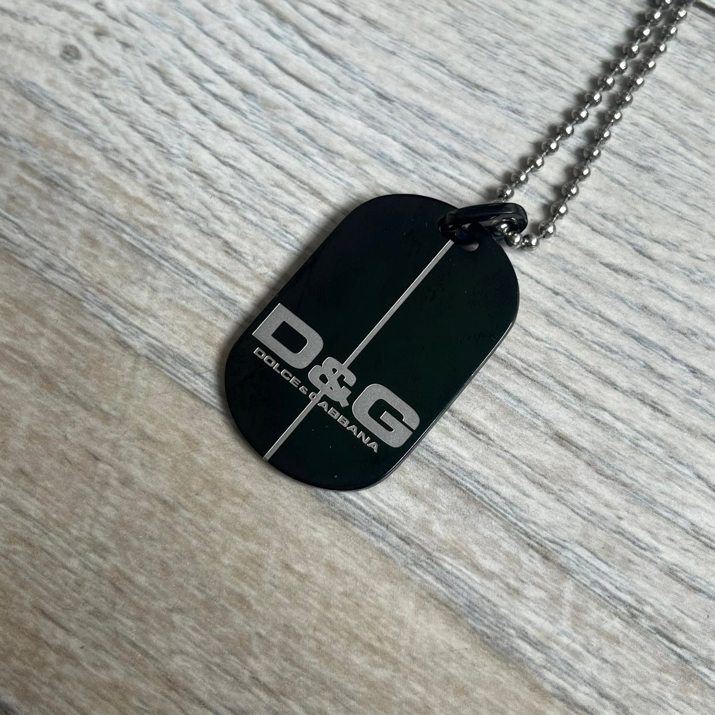 Dolce & Gabbana 2000s Split Black Dog Tag Necklace is a stainless steel necklace featuring a black dog tag pendant with a vertical grey split line running from top to bottom. The lower section of the pendant includes D&G and Dolce & Gabbana logos in the same grey tone as the split detail. The pendant has an iridescent black finish that shifts color depending on the angle. It is attached to a steel ball chain with a lobster clasp closure.