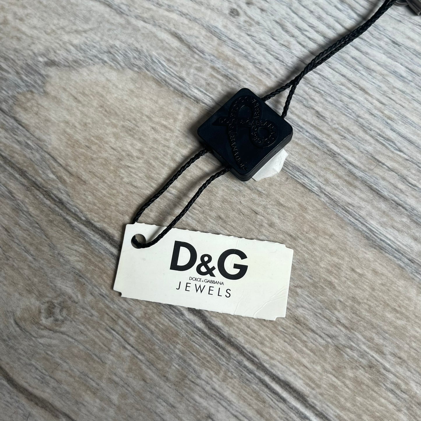 Dolce & Gabbana 2000s Split Black Dog Tag Necklace is a stainless steel necklace featuring a black dog tag pendant with a vertical grey split line running from top to bottom. The lower section of the pendant includes D&G and Dolce & Gabbana logos in the same grey tone as the split detail. The pendant has an iridescent black finish that shifts color depending on the angle. It is attached to a steel ball chain with a lobster clasp closure.