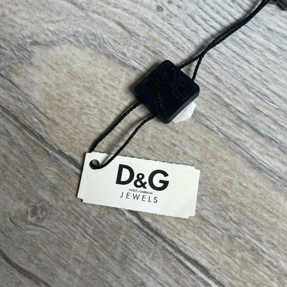 Dolce & Gabbana 2000s Split Black Dog Tag Necklace is a stainless steel necklace featuring a black dog tag pendant with a vertical grey split line running from top to bottom. The lower section of the pendant includes D&G and Dolce & Gabbana logos in the same grey tone as the split detail. The pendant has an iridescent black finish that shifts color depending on the angle. It is attached to a steel ball chain with a lobster clasp closure.