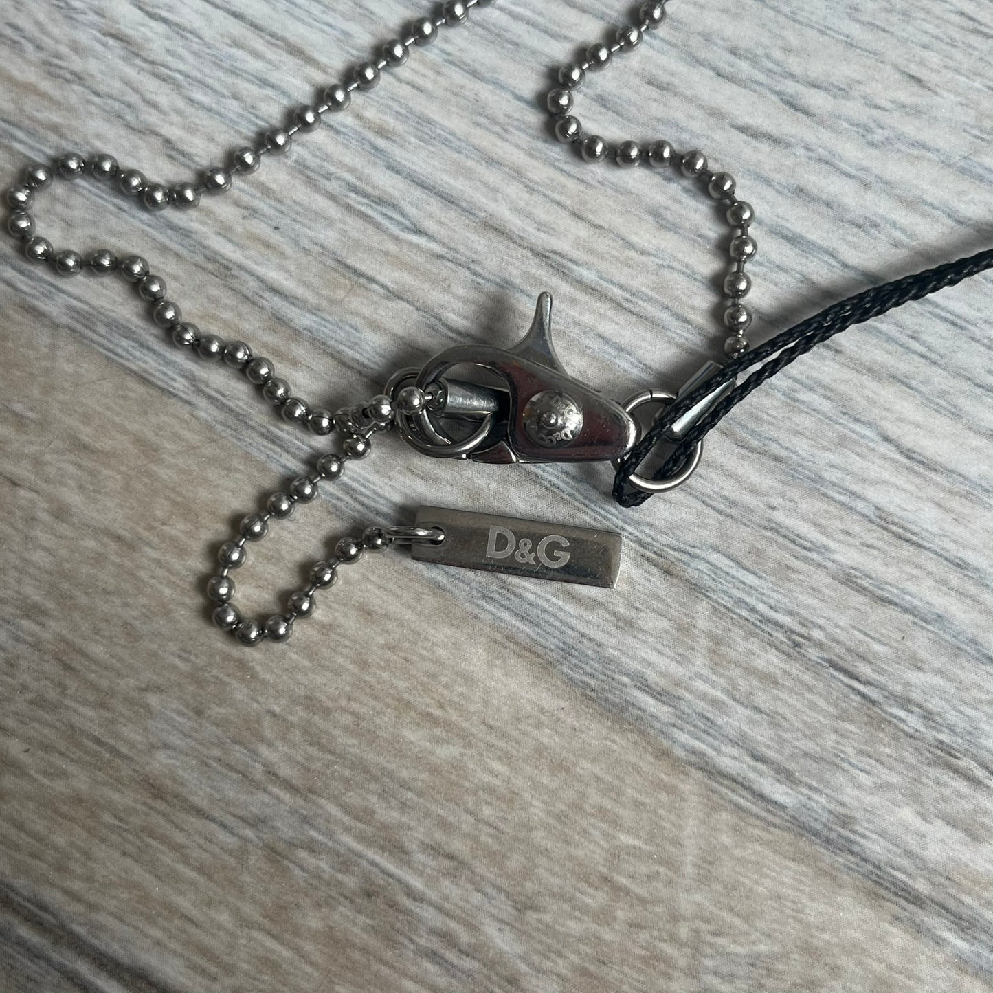Dolce & Gabbana 2000s Split Black Dog Tag Necklace is a stainless steel necklace featuring a black dog tag pendant with a vertical grey split line running from top to bottom. The lower section of the pendant includes D&G and Dolce & Gabbana logos in the same grey tone as the split detail. The pendant has an iridescent black finish that shifts color depending on the angle. It is attached to a steel ball chain with a lobster clasp closure.