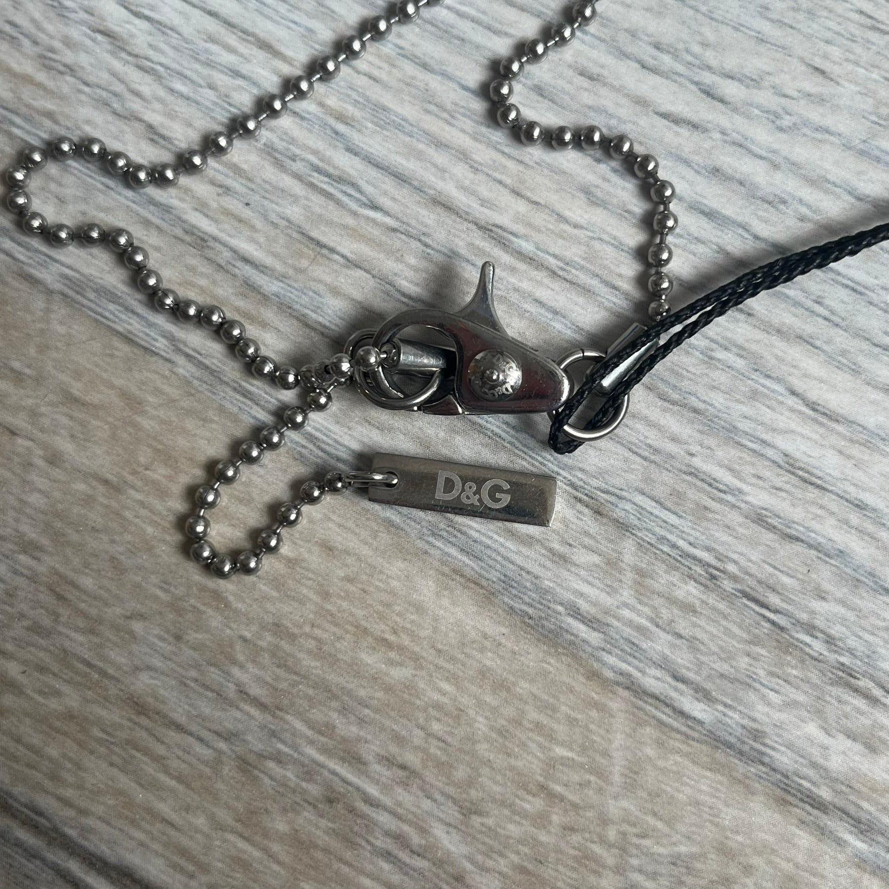 Dolce & Gabbana 2000s Split Black Dog Tag Necklace is a stainless steel necklace featuring a black dog tag pendant with a vertical grey split line running from top to bottom. The lower section of the pendant includes D&G and Dolce & Gabbana logos in the same grey tone as the split detail. The pendant has an iridescent black finish that shifts color depending on the angle. It is attached to a steel ball chain with a lobster clasp closure.
