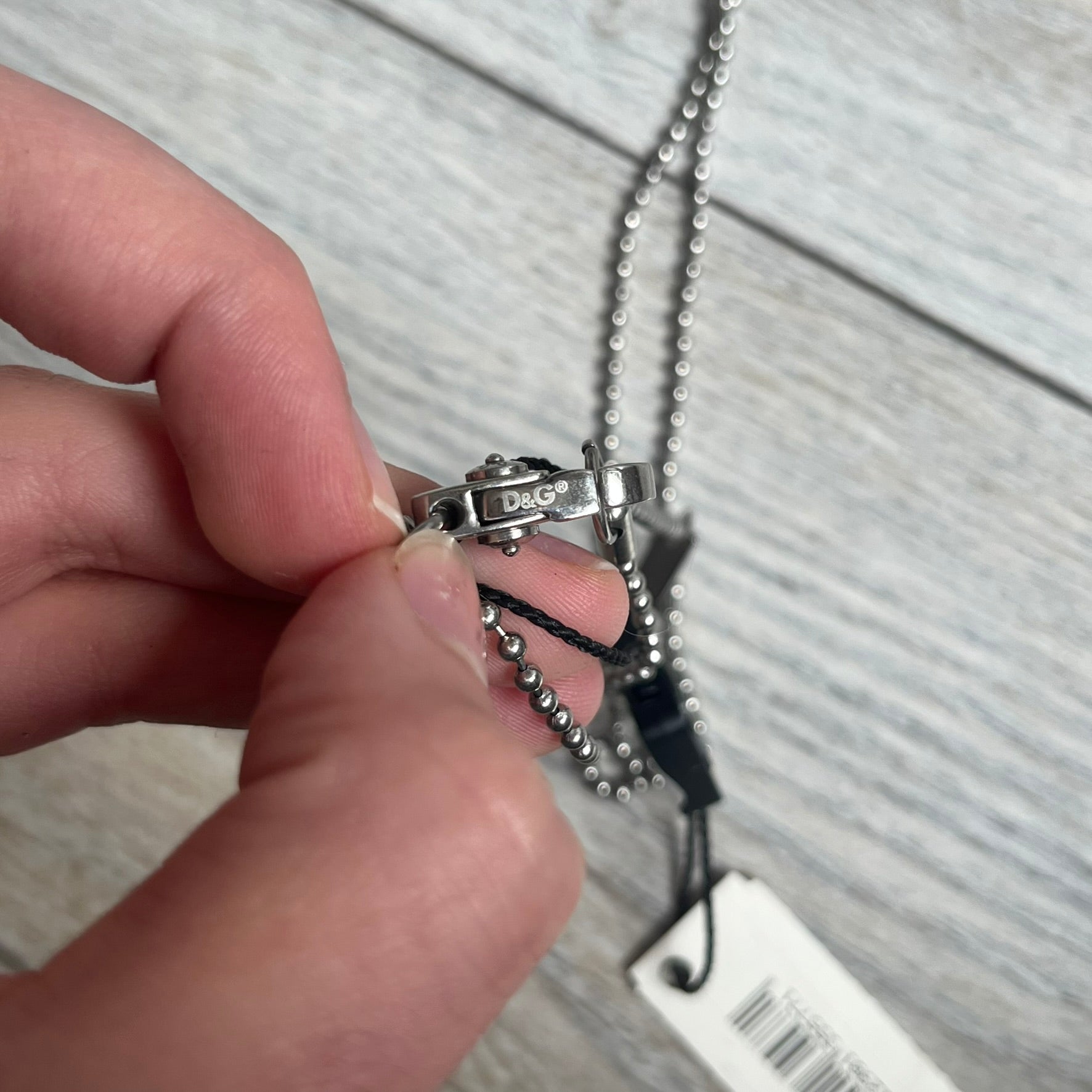 Dolce & Gabbana 2000s Split Black Dog Tag Necklace is a stainless steel necklace featuring a black dog tag pendant with a vertical grey split line running from top to bottom. The lower section of the pendant includes D&G and Dolce & Gabbana logos in the same grey tone as the split detail. The pendant has an iridescent black finish that shifts color depending on the angle. It is attached to a steel ball chain with a lobster clasp closure.