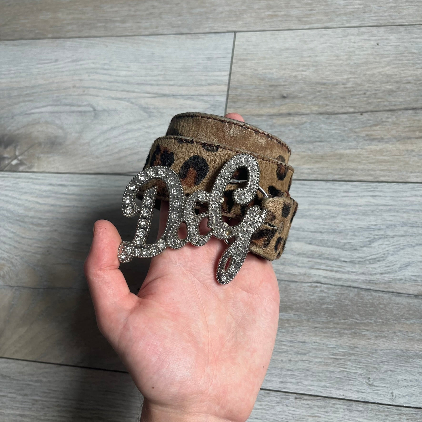 Dolce & Gabbana 2000s Studded Buckle Leopard Print Belt is a beige belt with an all-over leopard print and a short hair-textured exterior. The buckle features an italic-style D&G logo in a silver-tone finish with rhinestone detailing. The interior is finished in brown leather.