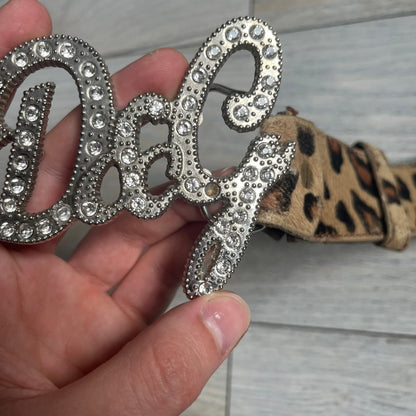 Dolce & Gabbana 2000s Studded Buckle Leopard Print Belt is a beige belt with an all-over leopard print and a short hair-textured exterior. The buckle features an italic-style D&G logo in a silver-tone finish with rhinestone detailing. The interior is finished in brown leather.