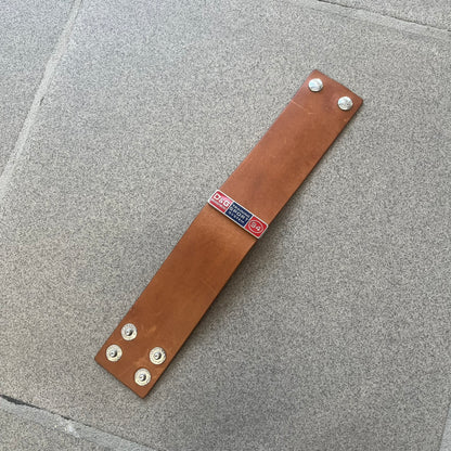 Dolce & Gabbana 2000s "Training Sport System" Bracelet (DJ0455) is a wide brown leather bracelet featuring a stainless steel plaque at the center. The plaque is detailed with a raised D&G logo, "Training Sport System" text, "94," and "3" graphics set against red and navy backgrounds. The interior is stamped with D&G branding and "Genuine Leather" markings. The bracelet fastens with two snap-button size adjustments and comes complete with its original box, dust bag, and papers.