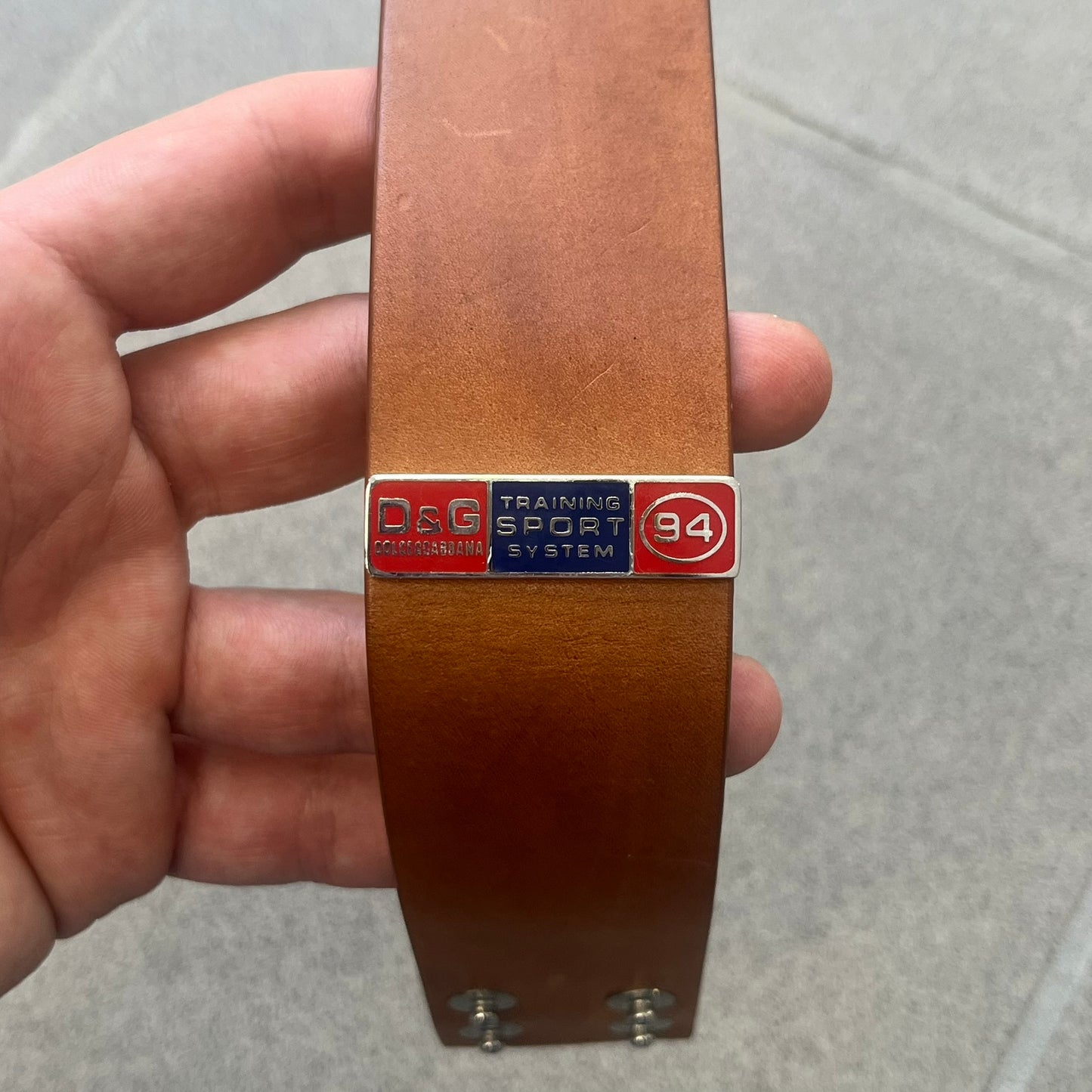 Dolce & Gabbana 2000s "Training Sport System" Bracelet (DJ0455) is a wide brown leather bracelet featuring a stainless steel plaque at the center. The plaque is detailed with a raised D&G logo, "Training Sport System" text, "94," and "3" graphics set against red and navy backgrounds. The interior is stamped with D&G branding and "Genuine Leather" markings. The bracelet fastens with two snap-button size adjustments and comes complete with its original box, dust bag, and papers.