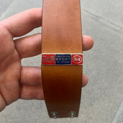 Dolce & Gabbana 2000s "Training Sport System" Bracelet (DJ0455) is a wide brown leather bracelet featuring a stainless steel plaque at the center. The plaque is detailed with a raised D&G logo, "Training Sport System" text, "94," and "3" graphics set against red and navy backgrounds. The interior is stamped with D&G branding and "Genuine Leather" markings. The bracelet fastens with two snap-button size adjustments and comes complete with its original box, dust bag, and papers.