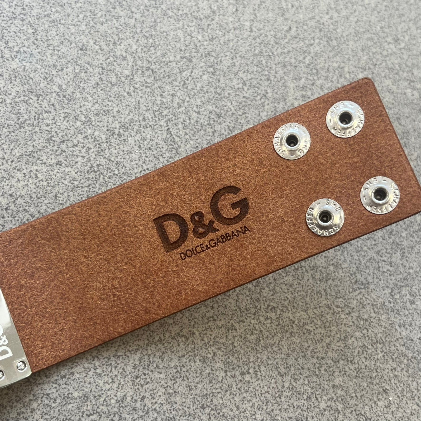 Dolce &amp; Gabbana 2000s "Training Sport System" Bracelet (DJ0455) is a wide brown leather bracelet featuring a stainless steel plaque at the center. The plaque is detailed with a raised D&amp;G logo, "Training Sport System" text and "94" graphics set against red and navy backgrounds. The interior is stamped with D&amp;G branding and "Genuine Leather" markings. The bracelet fastens with two snap-button size adjustments and comes complete with its original box, dust bag, and papers.