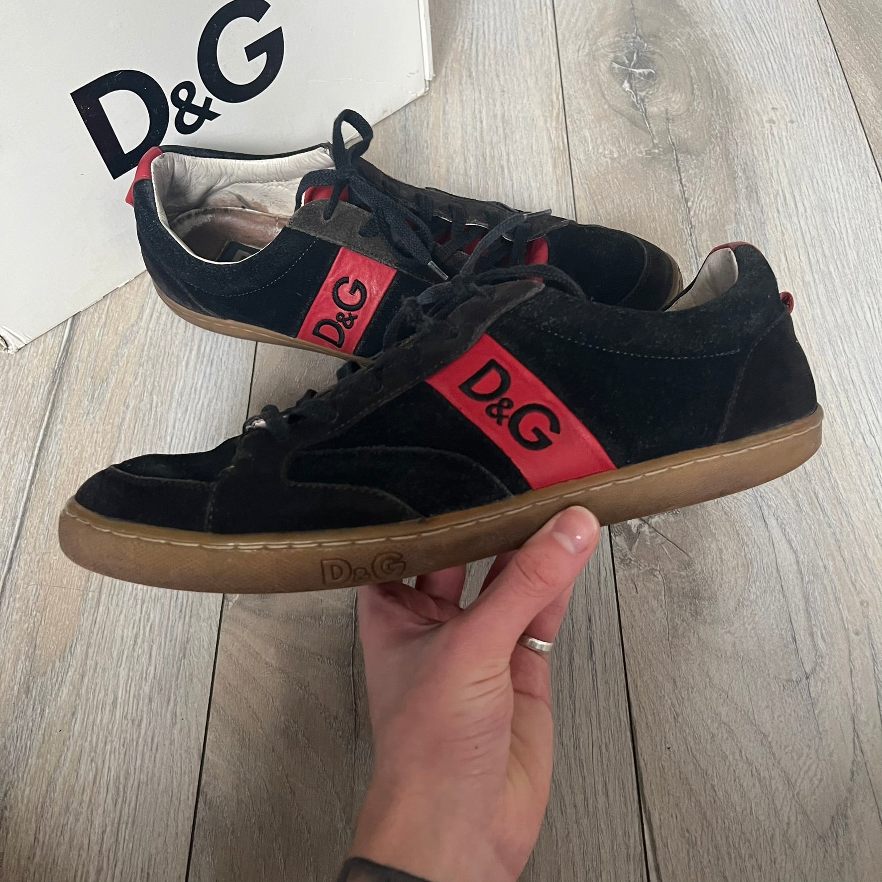 Dolce & Gabbana 2013 Bicolor Sneakers are black leather sneakers with a brown rubber sole. They feature red leather stripes on the sides, tongue details, and a back stripe, with black D&G logos on the sides and tongue. The sneakers come with the original box and tags.
