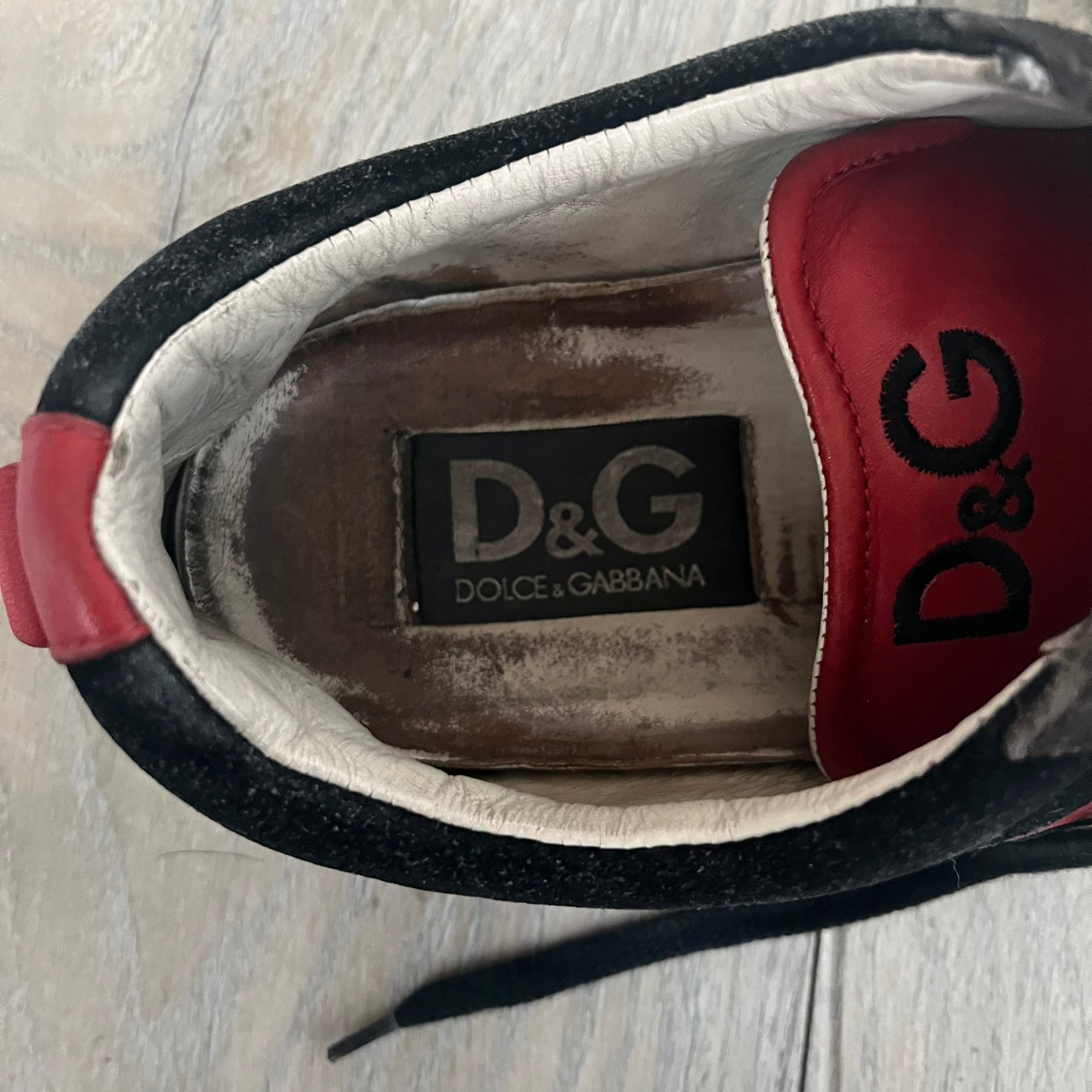 Dolce & Gabbana 2013 Bicolor Sneakers are black leather sneakers with a brown rubber sole. They feature red leather stripes on the sides, tongue details, and a back stripe, with black D&G logos on the sides and tongue. The sneakers come with the original box and tags.