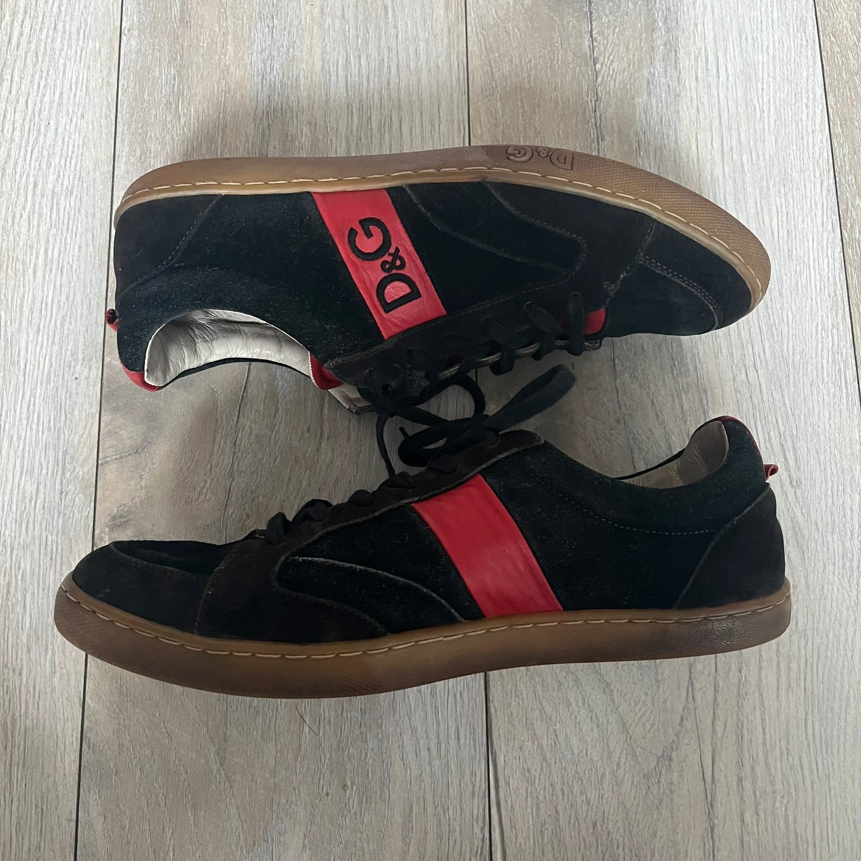 Dolce & Gabbana 2013 Bicolor Sneakers are black leather sneakers with a brown rubber sole. They feature red leather stripes on the sides, tongue details, and a back stripe, with black D&G logos on the sides and tongue. The sneakers come with the original box and tags.