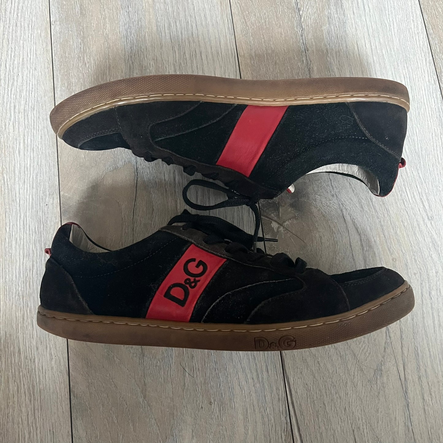 Dolce & Gabbana 2013 Bicolor Sneakers are black leather sneakers with a brown rubber sole. They feature red leather stripes on the sides, tongue details, and a back stripe, with black D&G logos on the sides and tongue. The sneakers come with the original box and tags.