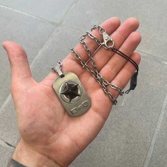 The Dolce &amp; Gabbana Star Dog Tag Necklace (DJ0889) is a heavy stainless steel necklace featuring a thick, oversized dog tag pendant. The front of the pendant has a raised black star mounted on a silver-tone circular base, with open cutout sections between the star and the circle. A raised D&amp;G logo is positioned at the lower front of the dog tag. The necklace is finished with a lobster clasp and has a solid, weighty construction throughout.