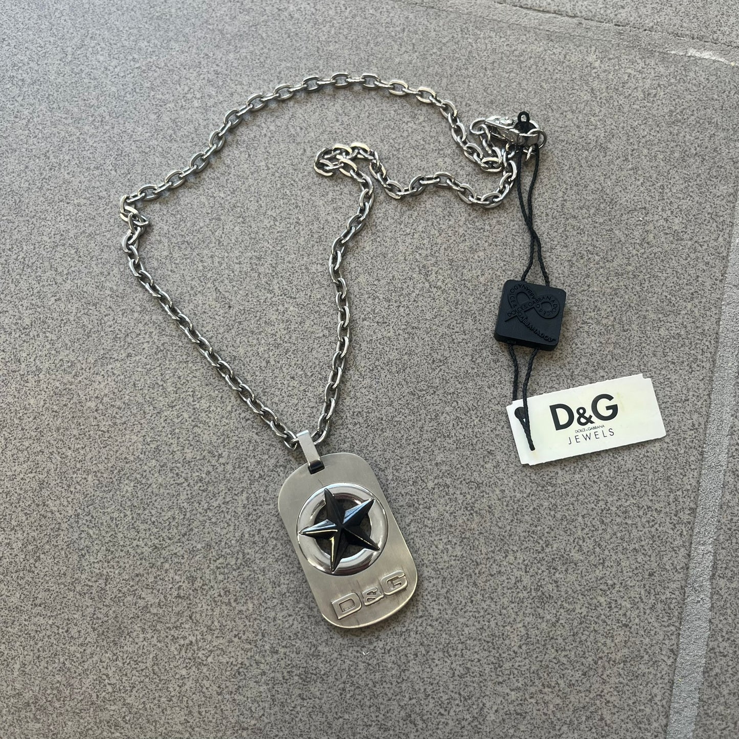 The Dolce &amp; Gabbana Star Dog Tag Necklace (DJ0889) is a heavy stainless steel necklace featuring a thick, oversized dog tag pendant. The front of the pendant has a raised black star mounted on a silver-tone circular base, with open cutout sections between the star and the circle. A raised D&amp;G logo is positioned at the lower front of the dog tag. The necklace is finished with a lobster clasp and has a solid, weighty construction throughout.