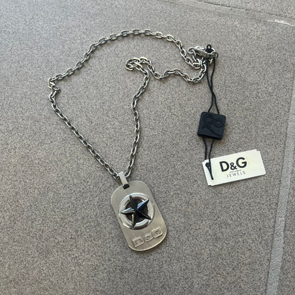 The Dolce &amp; Gabbana Star Dog Tag Necklace (DJ0889) is a heavy stainless steel necklace featuring a thick, oversized dog tag pendant. The front of the pendant has a raised black star mounted on a silver-tone circular base, with open cutout sections between the star and the circle. A raised D&amp;G logo is positioned at the lower front of the dog tag. The necklace is finished with a lobster clasp and has a solid, weighty construction throughout.