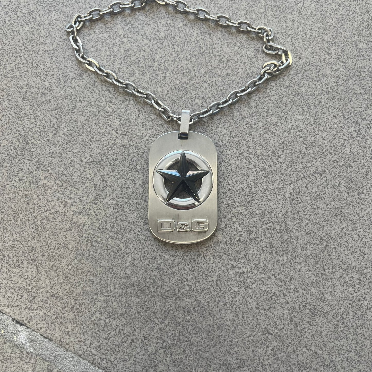 The Dolce &amp; Gabbana Star Dog Tag Necklace (DJ0889) is a heavy stainless steel necklace featuring a thick, oversized dog tag pendant. The front of the pendant has a raised black star mounted on a silver-tone circular base, with open cutout sections between the star and the circle. A raised D&amp;G logo is positioned at the lower front of the dog tag. The necklace is finished with a lobster clasp and has a solid, weighty construction throughout.