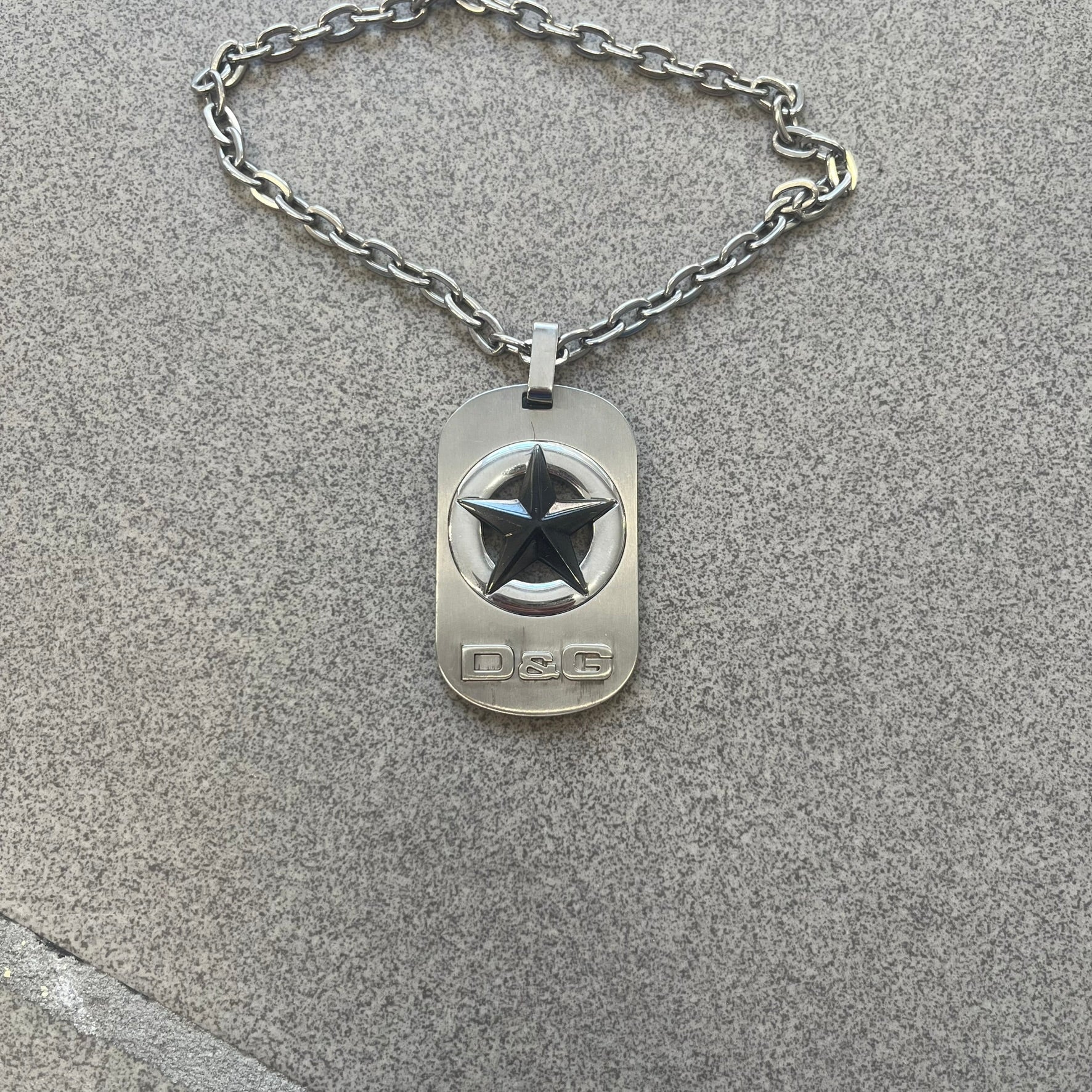 The Dolce &amp; Gabbana Star Dog Tag Necklace (DJ0889) is a heavy stainless steel necklace featuring a thick, oversized dog tag pendant. The front of the pendant has a raised black star mounted on a silver-tone circular base, with open cutout sections between the star and the circle. A raised D&amp;G logo is positioned at the lower front of the dog tag. The necklace is finished with a lobster clasp and has a solid, weighty construction throughout.