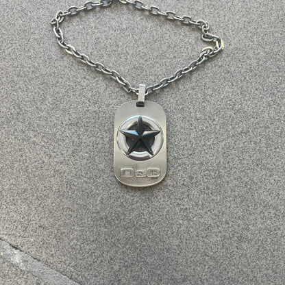 The Dolce &amp; Gabbana Star Dog Tag Necklace (DJ0889) is a heavy stainless steel necklace featuring a thick, oversized dog tag pendant. The front of the pendant has a raised black star mounted on a silver-tone circular base, with open cutout sections between the star and the circle. A raised D&amp;G logo is positioned at the lower front of the dog tag. The necklace is finished with a lobster clasp and has a solid, weighty construction throughout.