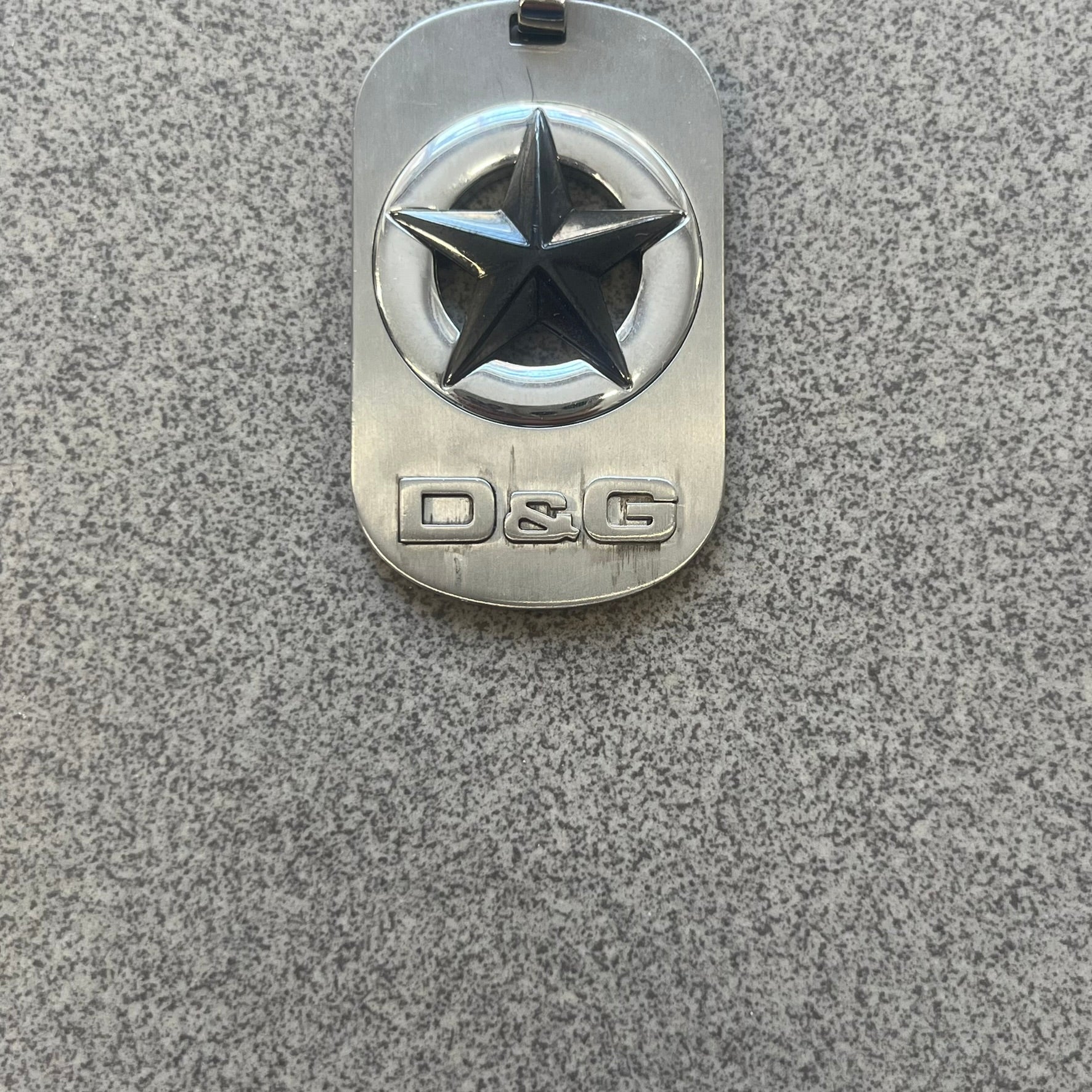 The Dolce &amp; Gabbana Star Dog Tag Necklace (DJ0889) is a heavy stainless steel necklace featuring a thick, oversized dog tag pendant. The front of the pendant has a raised black star mounted on a silver-tone circular base, with open cutout sections between the star and the circle. A raised D&amp;G logo is positioned at the lower front of the dog tag. The necklace is finished with a lobster clasp and has a solid, weighty construction throughout.