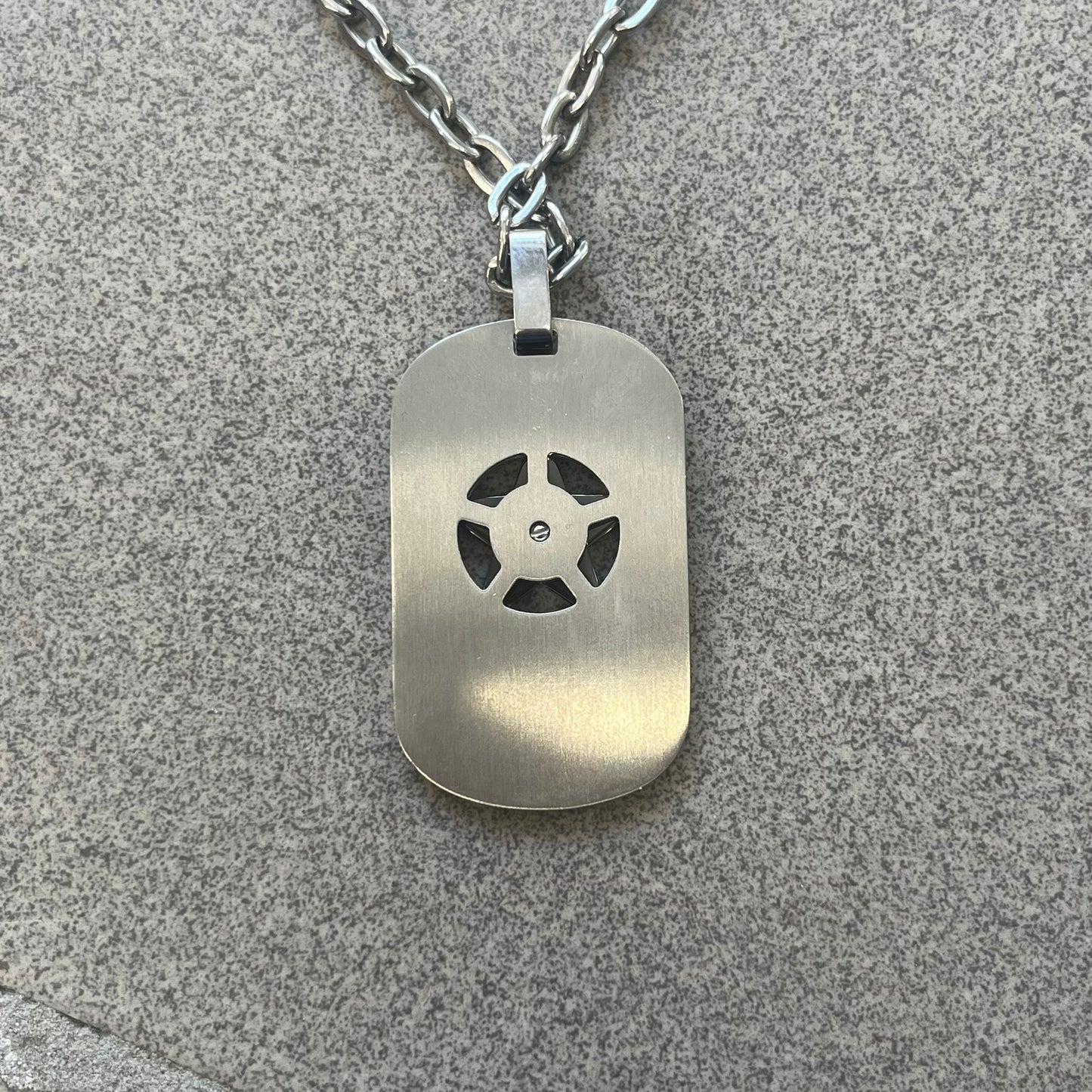 The Dolce &amp; Gabbana Star Dog Tag Necklace (DJ0889) is a heavy stainless steel necklace featuring a thick, oversized dog tag pendant. The front of the pendant has a raised black star mounted on a silver-tone circular base, with open cutout sections between the star and the circle. A raised D&amp;G logo is positioned at the lower front of the dog tag. The necklace is finished with a lobster clasp and has a solid, weighty construction throughout.