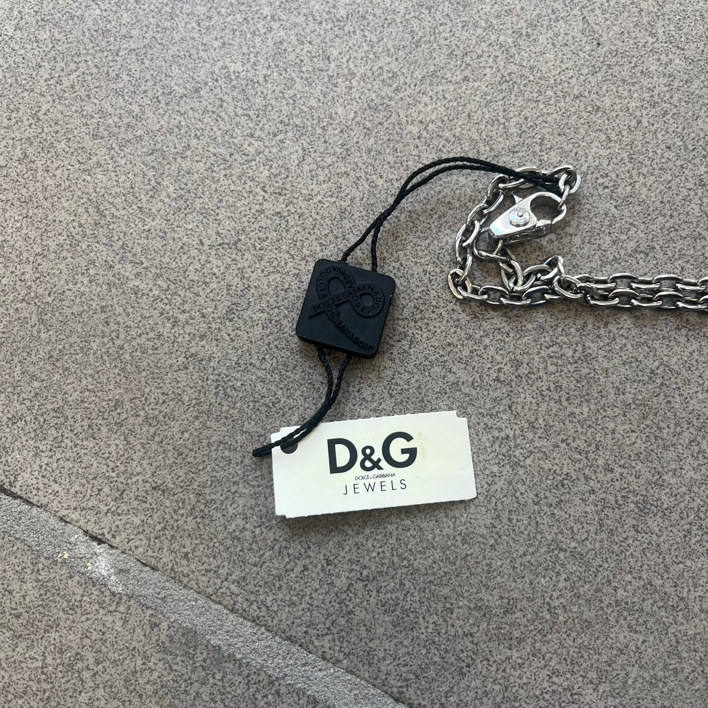 The Dolce &amp; Gabbana Star Dog Tag Necklace (DJ0889) is a heavy stainless steel necklace featuring a thick, oversized dog tag pendant. The front of the pendant has a raised black star mounted on a silver-tone circular base, with open cutout sections between the star and the circle. A raised D&amp;G logo is positioned at the lower front of the dog tag. The necklace is finished with a lobster clasp and has a solid, weighty construction throughout.