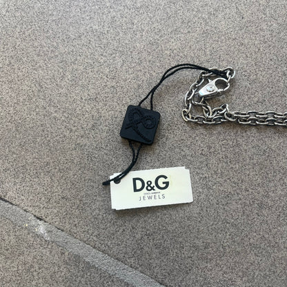 The Dolce &amp; Gabbana Star Dog Tag Necklace (DJ0889) is a heavy stainless steel necklace featuring a thick, oversized dog tag pendant. The front of the pendant has a raised black star mounted on a silver-tone circular base, with open cutout sections between the star and the circle. A raised D&amp;G logo is positioned at the lower front of the dog tag. The necklace is finished with a lobster clasp and has a solid, weighty construction throughout.