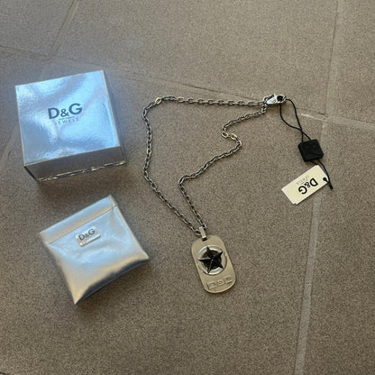 The Dolce &amp; Gabbana Star Dog Tag Necklace (DJ0889) is a heavy stainless steel necklace featuring a thick, oversized dog tag pendant. The front of the pendant has a raised black star mounted on a silver-tone circular base, with open cutout sections between the star and the circle. A raised D&amp;G logo is positioned at the lower front of the dog tag. The necklace is finished with a lobster clasp and has a solid, weighty construction throughout.