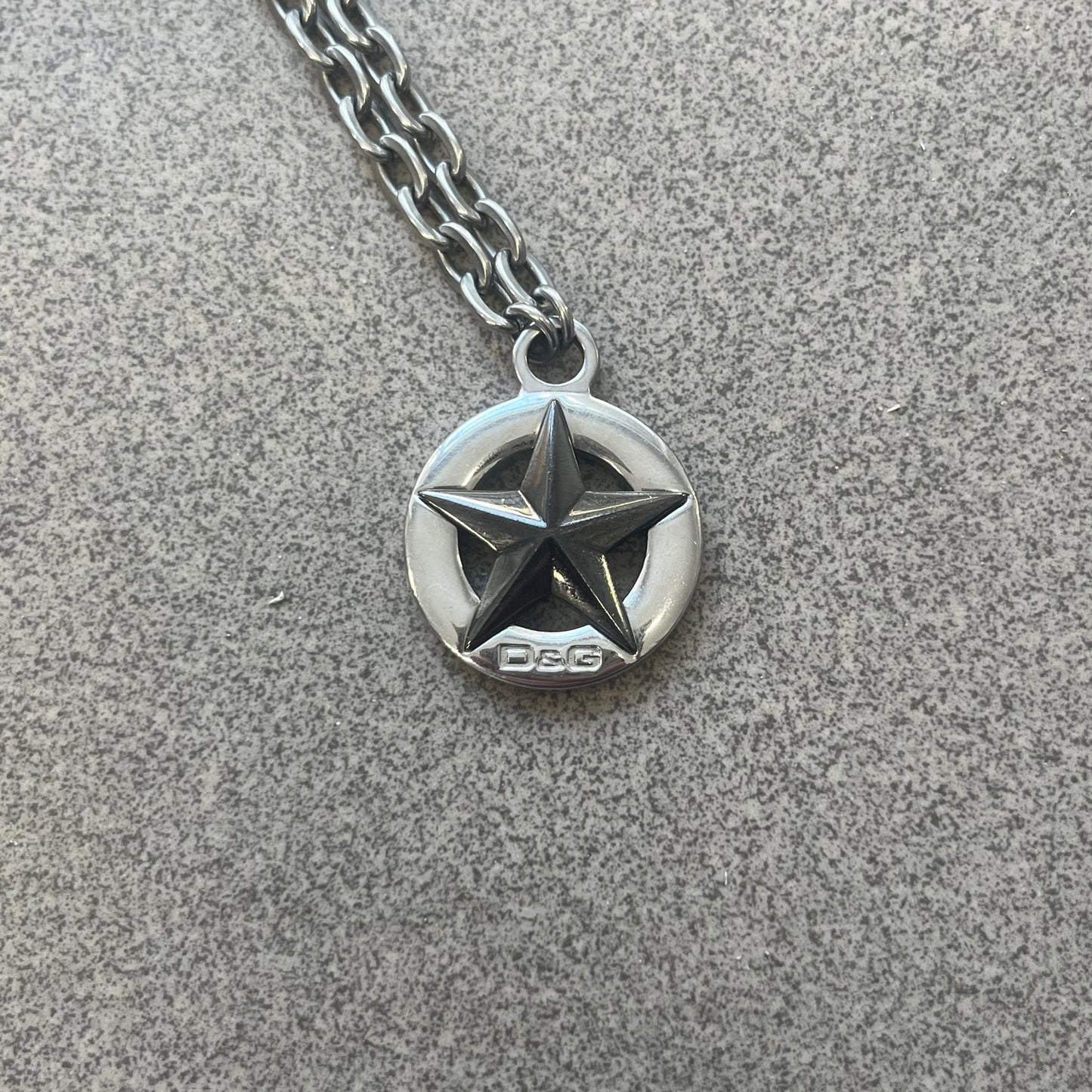 Dolce &amp; Gabbana AW2009 Military Star Necklace in grey. Features a 3D black star mounted on a silver-tone metal circle with an engraved D&amp;G logo at the bottom, suspended from a silver cable chain. Lobster clasp with small D&amp;G markings.