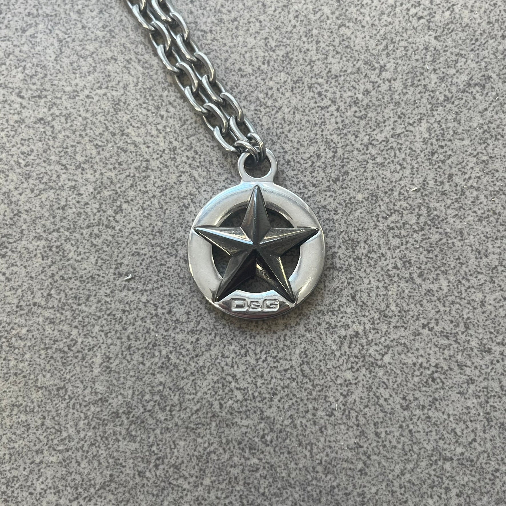 Dolce &amp; Gabbana AW2009 Military Star Necklace in grey. Features a 3D black star mounted on a silver-tone metal circle with an engraved D&amp;G logo at the bottom, suspended from a silver cable chain. Lobster clasp with small D&amp;G markings.