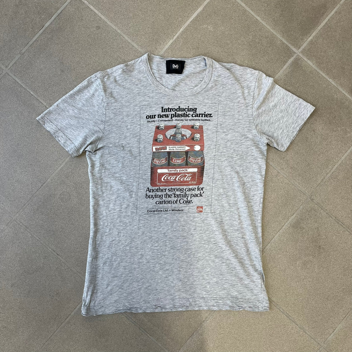 Dolce & Gabbana AW2011 Coca Cola Tee is a gray T-shirt made in collaboration with Coca-Cola, featuring a vintage promo-style graphic on the front of a six-pack of Coca-Cola bottles with “Introducing our new plastic carrier” text. It has licensed tags and a small black D&G logo on the upper back.