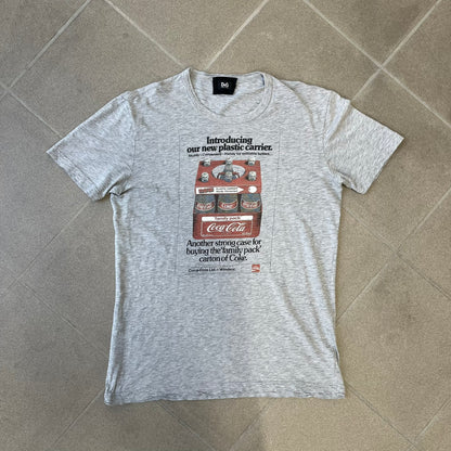 Dolce & Gabbana AW2011 Coca Cola Tee is a gray T-shirt made in collaboration with Coca-Cola, featuring a vintage promo-style graphic on the front of a six-pack of Coca-Cola bottles with “Introducing our new plastic carrier” text. It has licensed tags and a small black D&G logo on the upper back.
